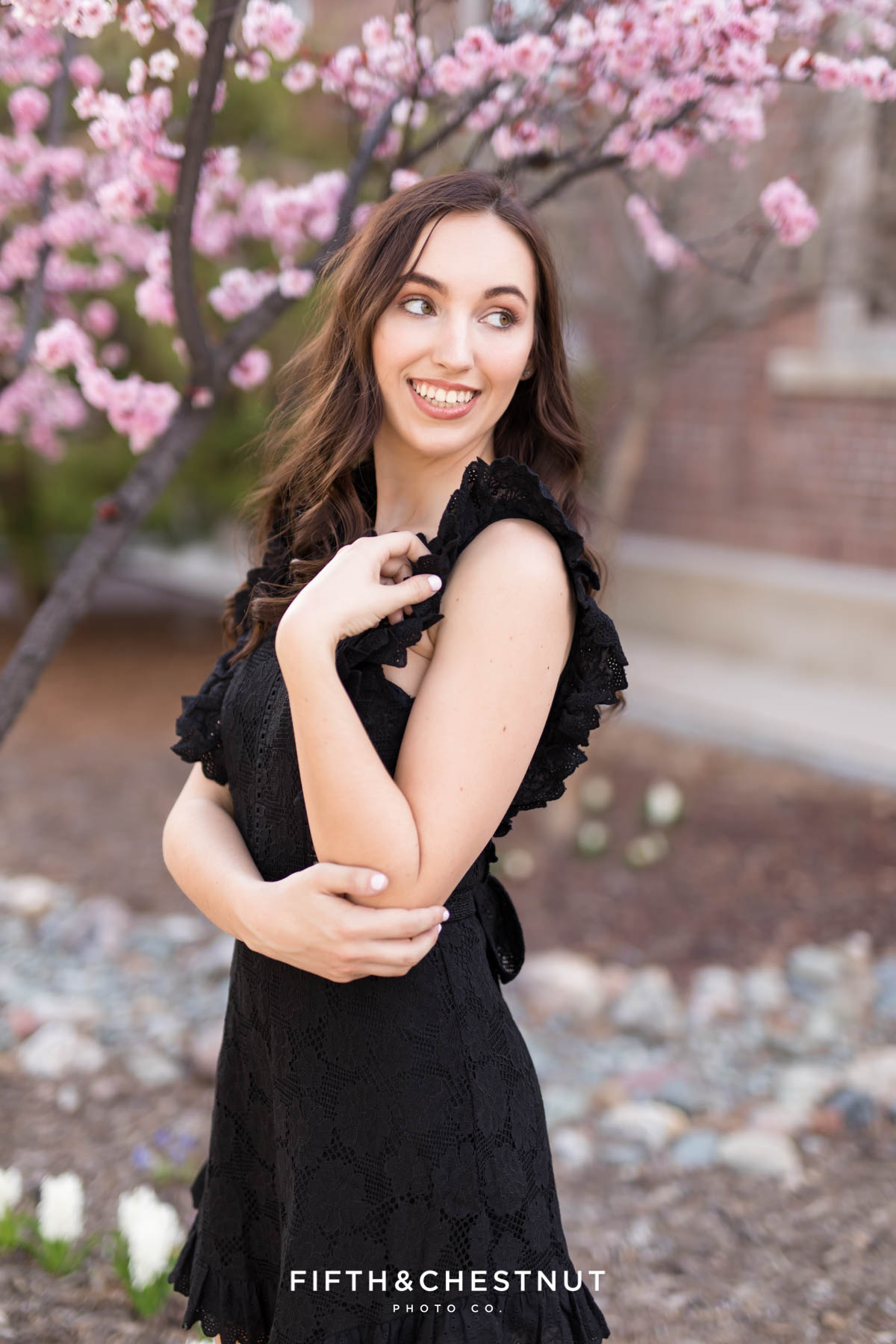 A unr graduate in a black dress stands in front of a pink blossoming plum tree for her spring UNR graduation portraits taken by Reno Graduation Photographer Fifth and Chestnut