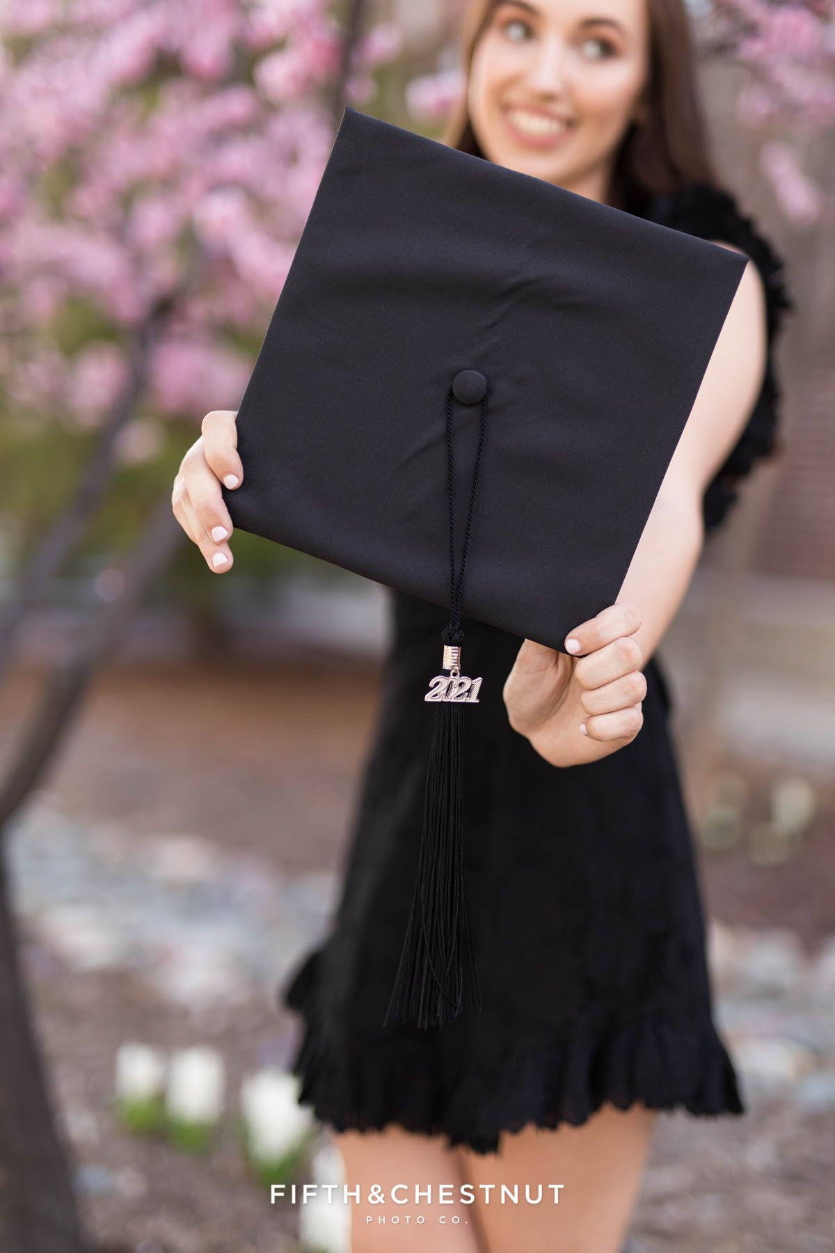 A UNR graduate holds her cap towards the camera to show off her 2021 tassel in front of an ornamental blossoming plum tree