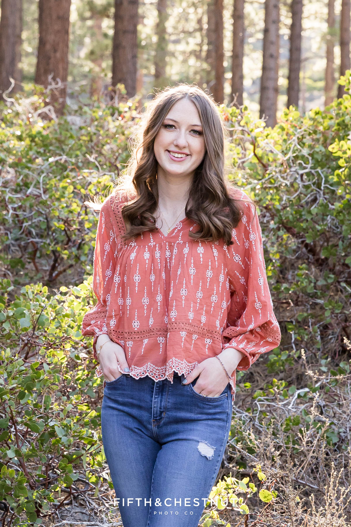 A reno student wearing a coral pink boho top has her hands in her pockets as she stands in front of manzanita for her forest high school senior portraits