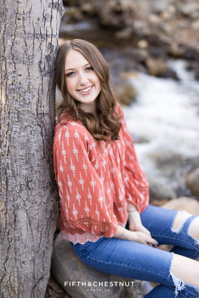 A Damonte Ranch High School Senior smiles for her forest high school senior portraits by a creek and a tree as an example of how to make your Reno/Tahoe senior portraits unique