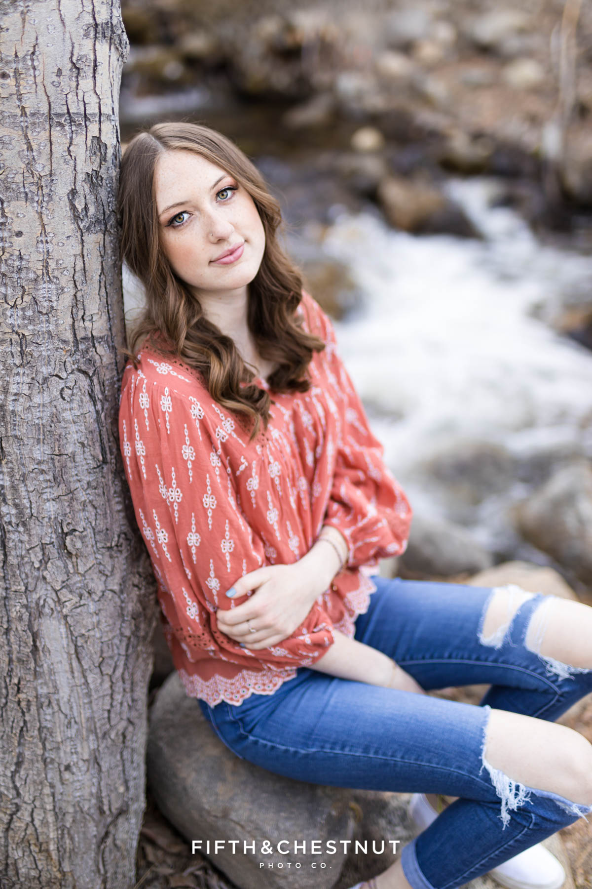 a student wearing coral pink leans against a tree by the creek for her reno senior pics