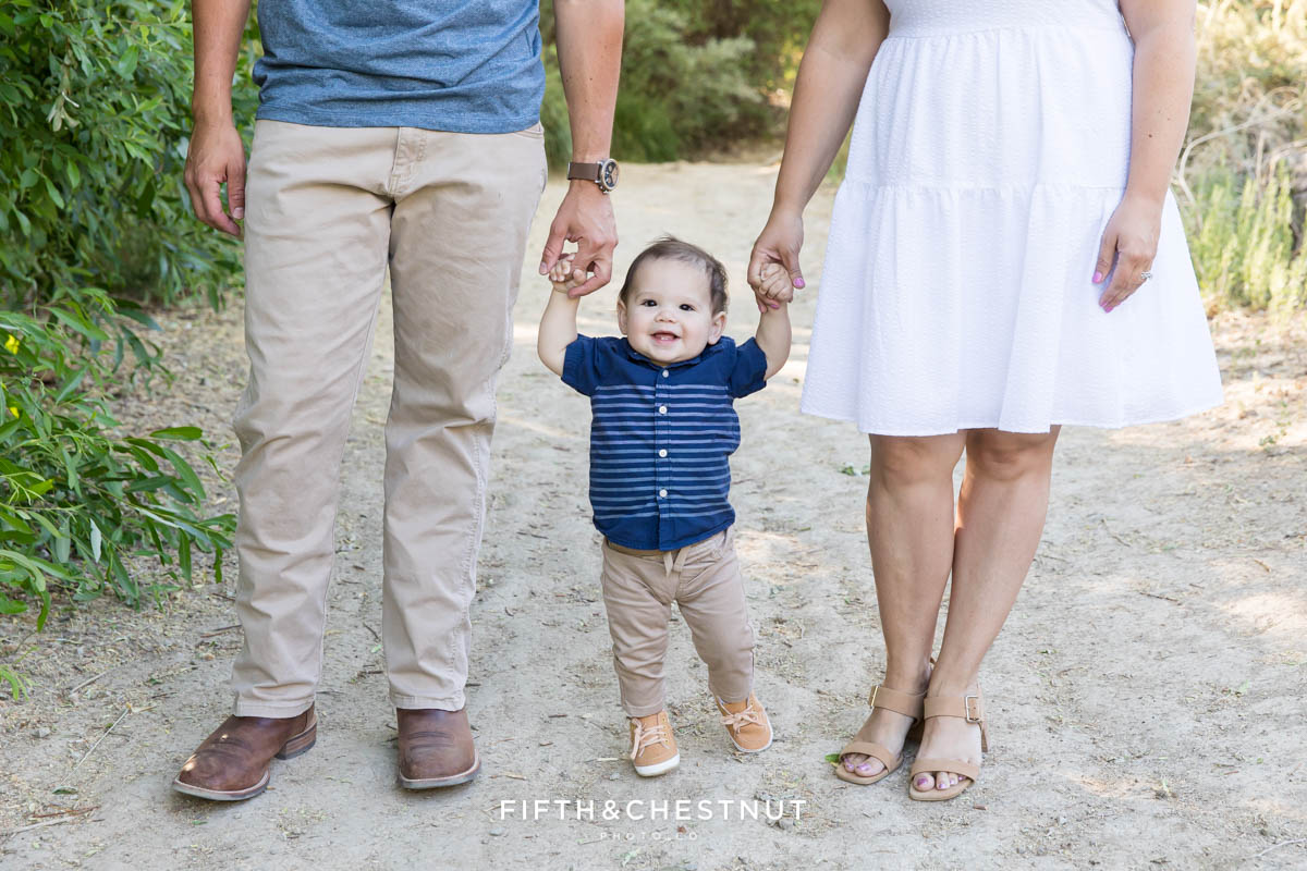 A one year old boy walks in between his parents as he holds their hands down the trail for their happy spring family photos