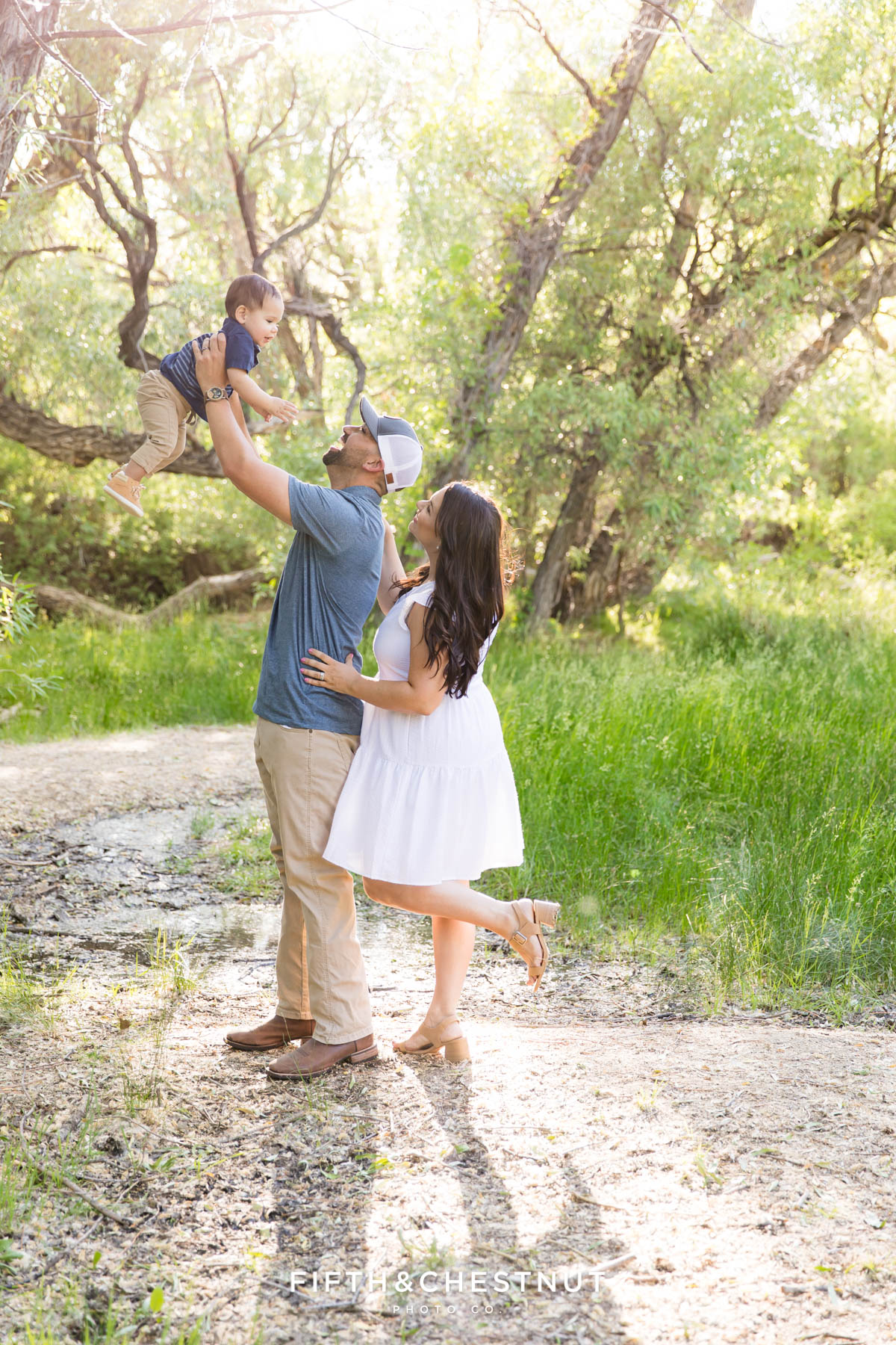 A dad holds up his one year old sone wearing a striped blue shirt with a meadow and trees behind them as his wife embraces him while wearing a white dress