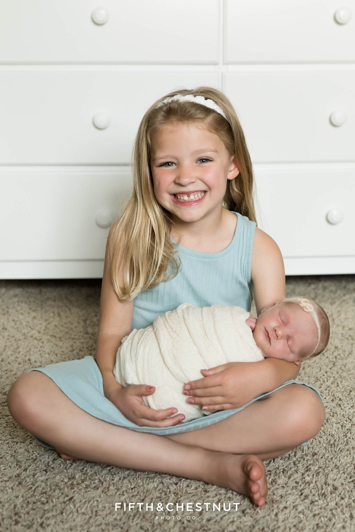 A happy big sister holds her newborn baby sister during her newborn twin photo shoot by Reno Family Photographer