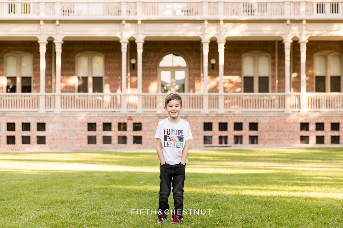 A 6 year old boy stands with his hands in his pockets wearing a shirt that says "Future Leader" in front of the Morrill Hall UNR alumni building on the south quad for child portraits by Reno Child Photographer