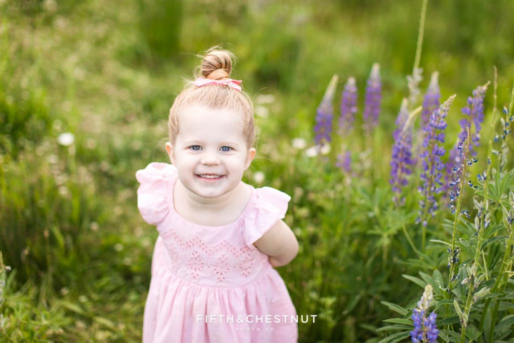 a three year old blonde girl in a pink dress smiles happily in a patch of wildflowers for her spring photoshoot by reno child photographer