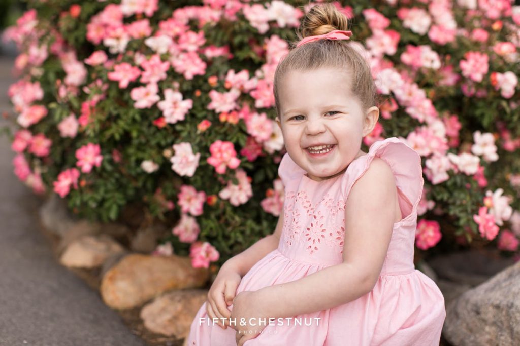 a sweet blonde three year old girl smiles in front of a gorgeous pink rose bush for spring wildflower portraits by reno child photographer