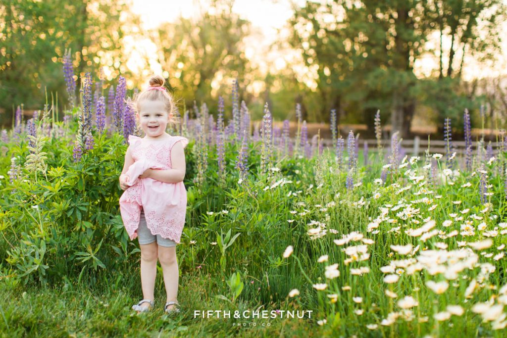 A blonde three year old girl in a pink dress stands in a wildflower patch full of lupines and daisies for spring wildflower portraits by reno child photographer