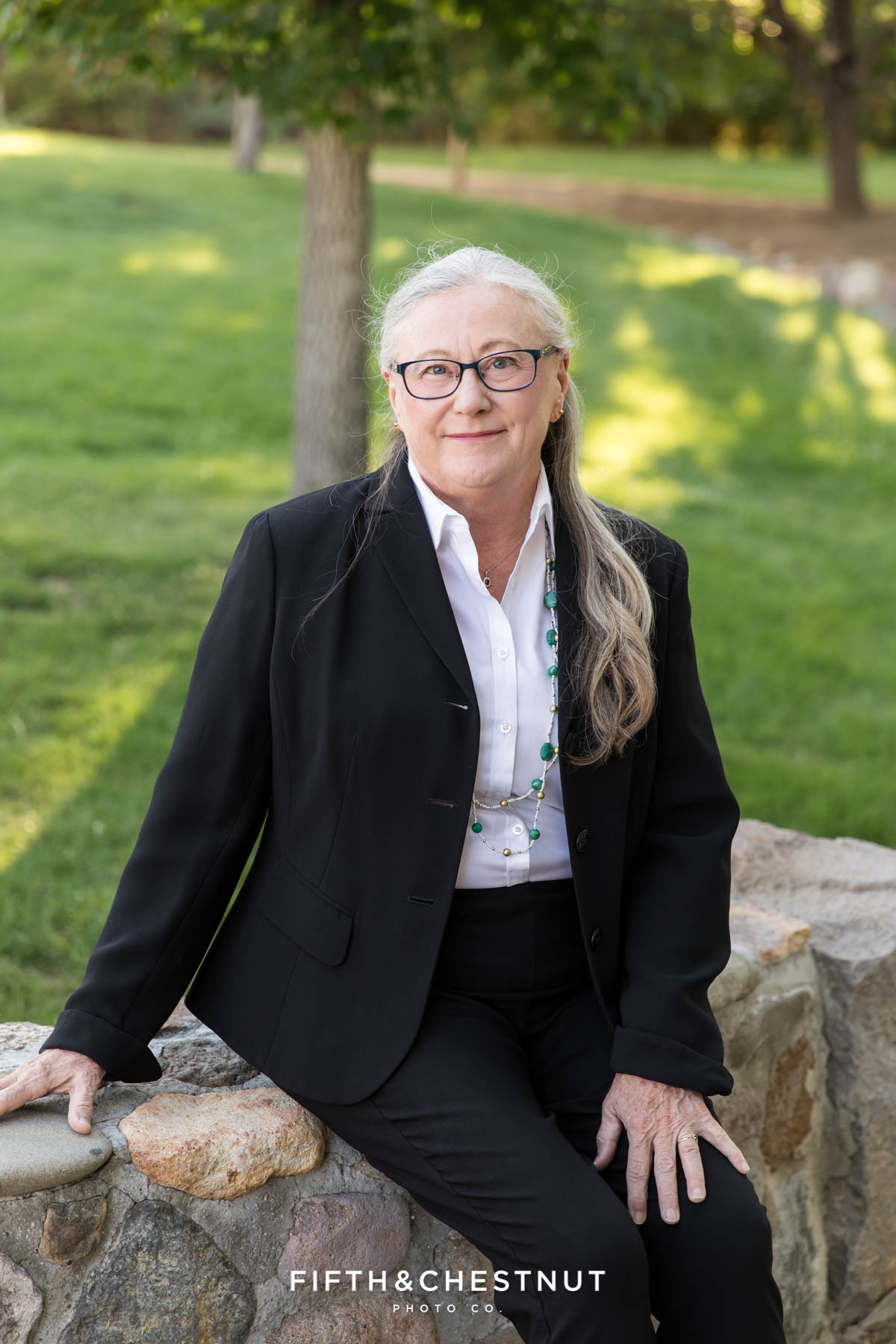woman sitting on a stone wall wearing a black suit jacket for corporate portraits in the park