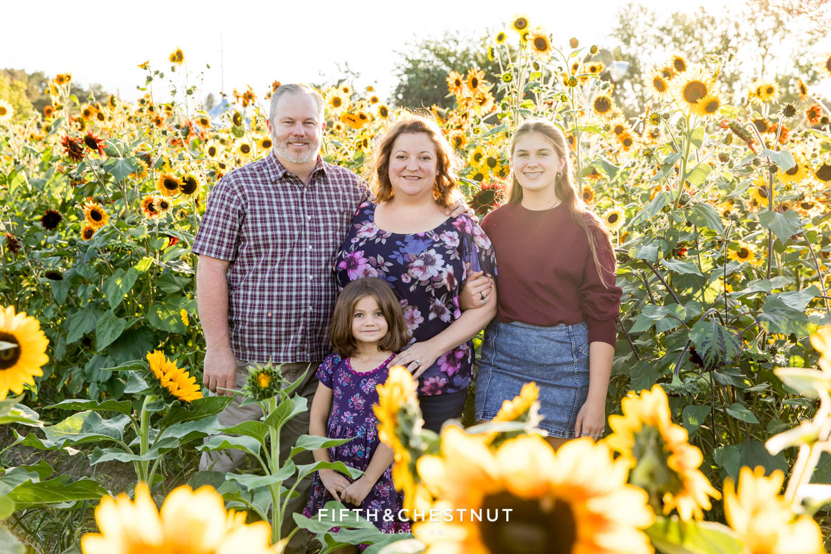 A family of four wearing tones of purple stands in a lush field of sunflowers as the golden sun sets behind them at Ferrari Farms for their sunflower family photos in summer.