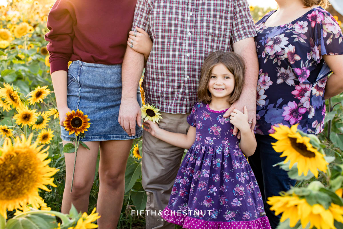 A family of four wearing tones of purple stands in a lush field of sunflowers as the golden sun sets behind them at Ferrari Farms for their sunflower family photos in summer.