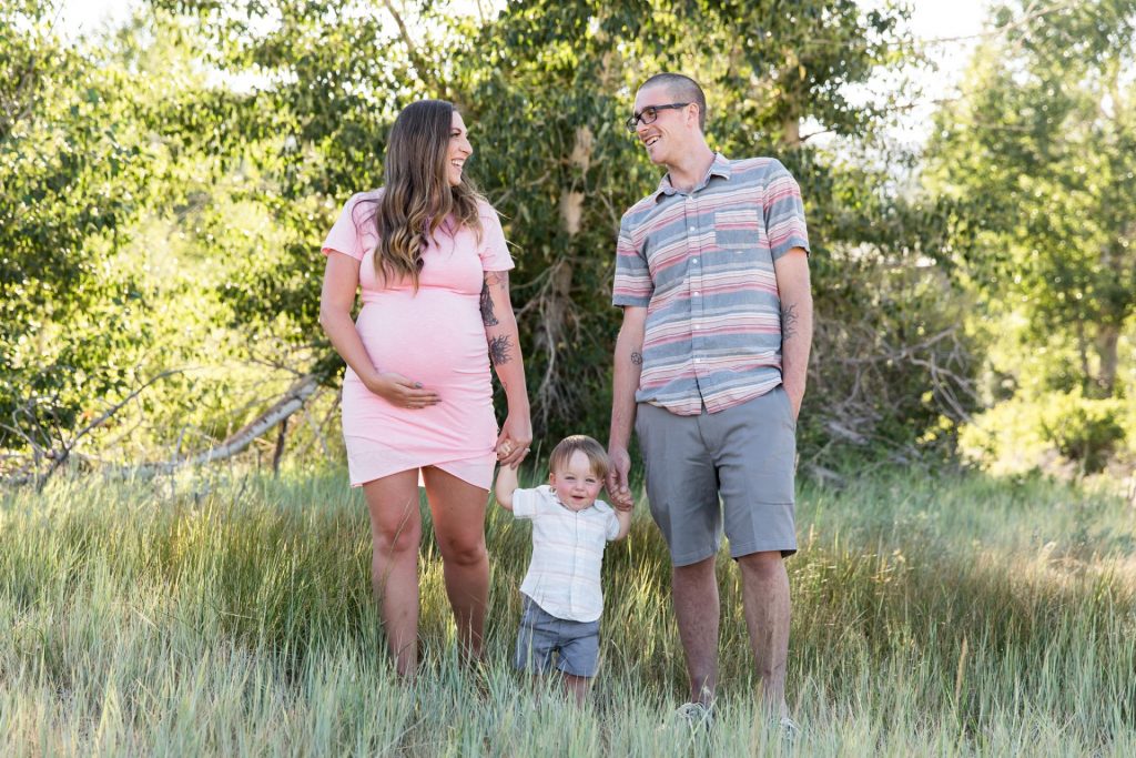 A lovely pregnant mom wearing a pink dress holds the hand of her sweet toddler who holds the hand of his dad in a meadow for reno family portraits by Reno Family Photographer