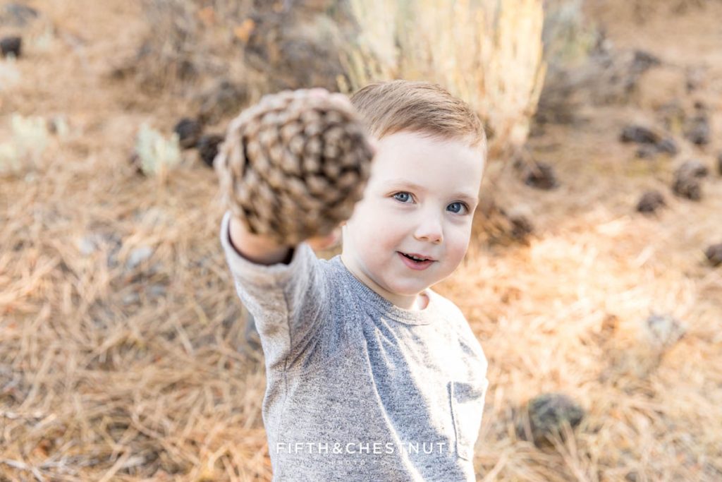 A young boy shows a pine cone to his photographer and smiles