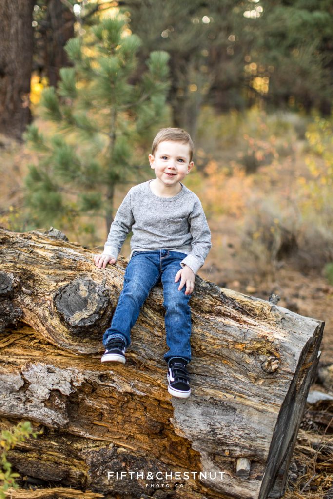 a four year old boy sitting on a log dangling his legs with fall foliage behind him