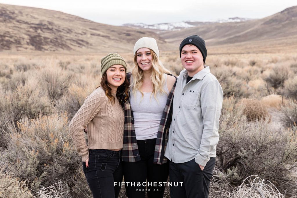 Three grown children huddle together for desert family portraits at Golden Eagle Regional Park wearing beanies for a photo by Reno Family Photographer