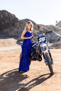 A teen in a sequin blue dress stands holding up a dirt bike in the desert for Moon Rocks senior photos