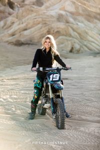 A blonde teen in dirt bike gear sits on her dirt bike with the sun radiating from behind her for Magical Moon Rocks Senior Portraits by Reno Senior Photographer