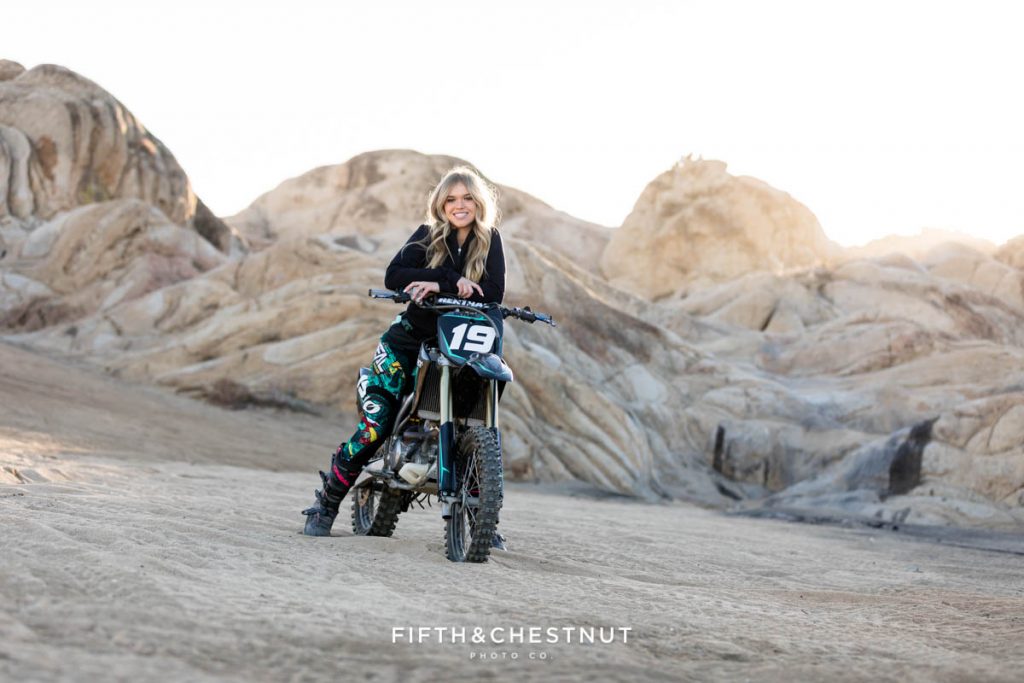 A female high school student dressed in brightly colored dirt bike attire poses with her black and teal dirt bike at Moon Rocks in Nevada for her unique Reno Senior Portraits by Reno Senior Photographer