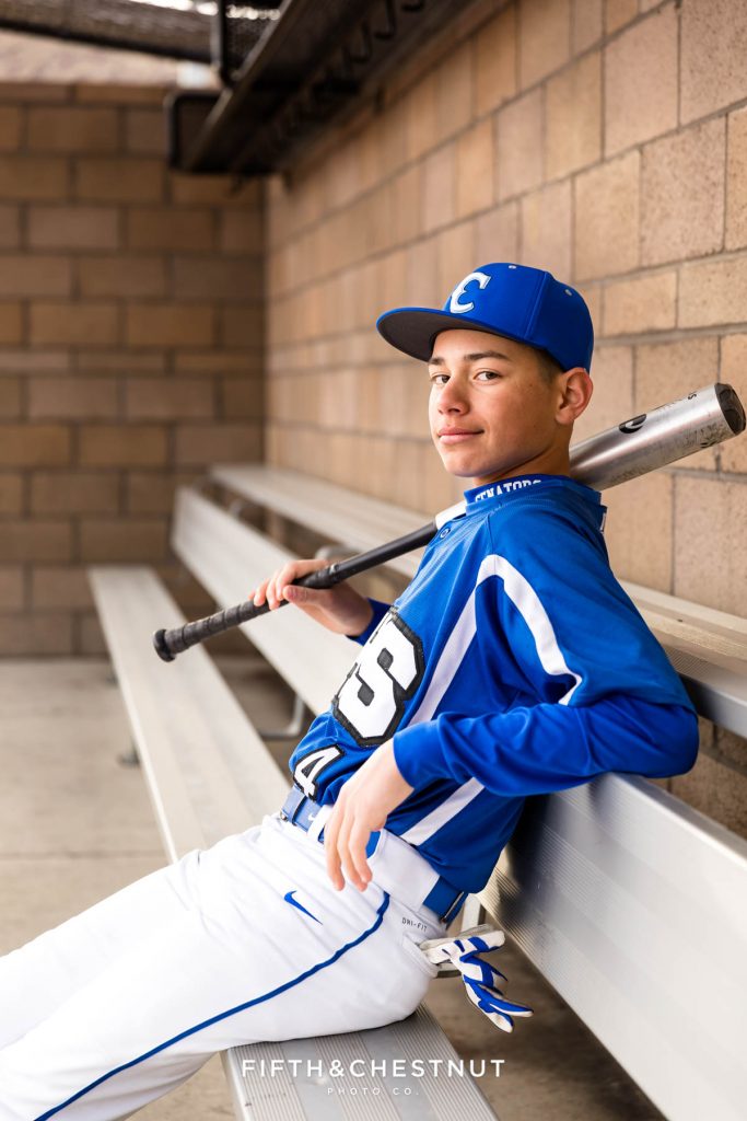 A baseball player rests in the dugout with his bat and baseball cap and gloves for his Reno senior portraits by Reno Senior Photographer