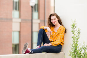 A Reno High School Senior girl wearing a floral yellow shirt, dark jeans and burgundy converse sneakers sits on the ledge of a retaining wall at UNR for her Reno Senior Portraits by Reno Senior Photographer
