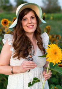 A red headed woman wearing a sun hat and holding a sunflower and garden shears in a white and floral ruffled shoulder dress in a field of sunflowers