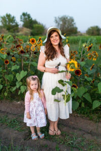 A red headed woman wearing a sun hat and holding a sunflower and garden shears in a white and floral ruffled shoulder dress in a field of sunflowers with a little blonde toddler wearing a floral dress and pink cardigan by her side