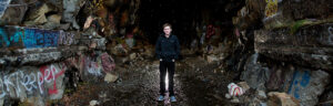 A Reno High School Senior boy stands in the middle of the Donner Summit Tunnels as water drips down behind him with a bright light shining from behind showcasing the graffiti on the boulders to his side