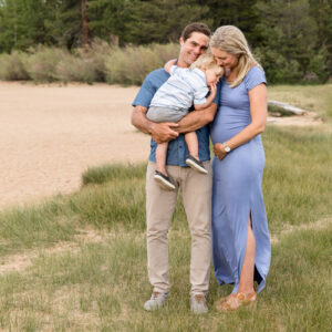 A family of three cuddles while standing in a beach meadow at Kiva Beach for their Lake Tahoe Family Portraits by Lake Tahoe Family Photographer on a cloudy summer evening