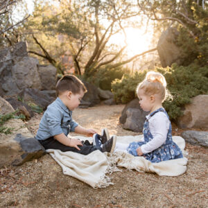 An adorable set of fraternal twins wearing denim outfits sits on a blanket and play together while the sun sets behind them in a high desert area surrounded by boulders and rocks during a child portrait session with Reno Family Photographer Fifth and Chestnut Photo Co.