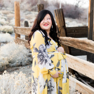 A pregnant woman with long black hair and red lips looks off into the distance as she wears a gorgeous yellow, white and gray maternity wrap dress for her maternity photos in reno by Reno Maternity Photographer