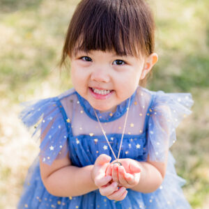 A sweet 3-year-old girl wearing a periwinkle dress with shiny star details holds her aquamarine necklace in her hand as she poses for her child portraits by Reno Child Photographer