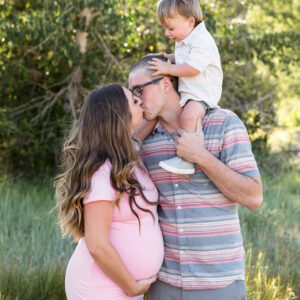 A pregnant couple wearing pink and gray clothing kisses while their son sits on his dad's shoulders for a fun and candid reno family photo by Reno Family Photographer