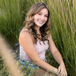 A Carson High School senior wearing a white, pink and green floral crop cami and jean shorts poses among tall grass for her senior portraits by Reno Senior Photographer on a warm summer evening