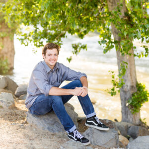 A Reno High School senior boy wearing jeans, a blue shirt and black hi-top vans sits on boulders by the Truckee River under a lush green tree for his Reno senior portraits by Reno's best high school senior photographer