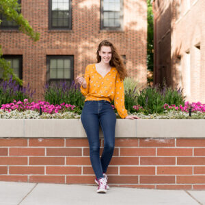 A McQueen high school senior sits against the brick retaining wall with a garden of purple and pink flowers behind her at UNR for her reno senior photography by Reno Senior Photographer