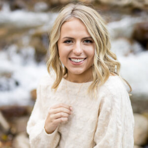 A Reno High School Senior plays with her gold necklace in front of a creek bed while wearing a cozy ivory sweater with soft tousled blonde hair for her Reno Senior photos by Reno Senior Photographer