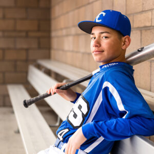 A Carson High School senior wearing his baseball uniform holds a bat in his right hand while also wearing his Carson High School baseball cap in the dugouts at Golden Eagle Regional Park and Sports Complex for his Carson Senior Portraits by Carson Senior Photographer