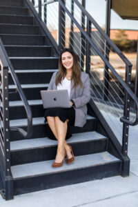 A brunette woman in a suit jacket, white shirt and black pencil skirt sits on the steps of REMAX with her MacBook Pro for her professional branding photos by Reno Headshot Photographer