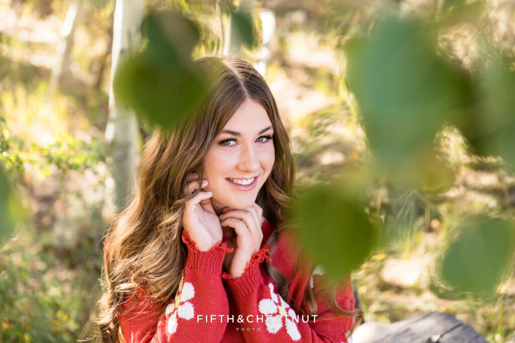 A senior portrait of a girl wearing a red sweater with daisy details, posing with her hands near her face with green aspen leaves in the foreground.