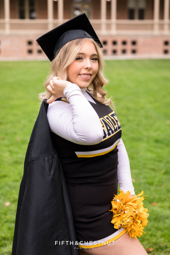 A Galena High School Senior poses on the UNR quad in her cheer uniform with gold pom poms wearing her graduation cap and hanging her grad gown over her shoulder as an example of why Senior photos are worth the cost