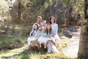 A family wearing light blue and white sits on a wooden stump and cuddles up for family portraits in Spring by Reno Family Photographer at Rancho San Rafael Regional Park in Reno, NV.