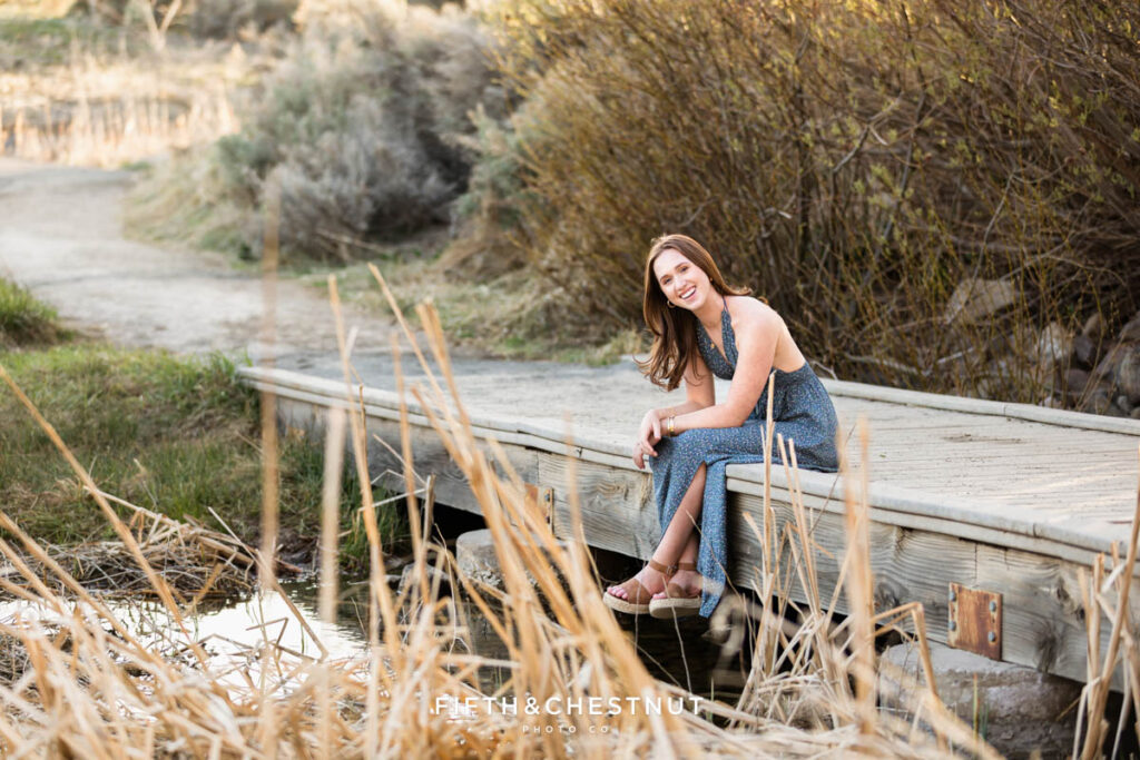 Luminous spring boho Reno senior portraits at Rancho San Rafael by Reno Senior Photographer with Ava in a floral blue maxi dress and sandals sitting on a wooden bridge by the pond with cattails in the foreground.