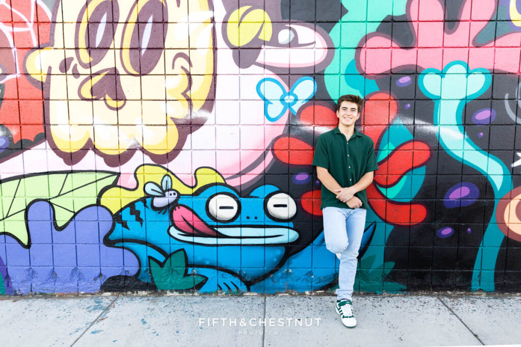 A senior wearing a casual green short sleeve button up and light washed jeans in front of a Hannah Eddy art installation in Midtown Reno for senior portraits by Reno Senior Photographer