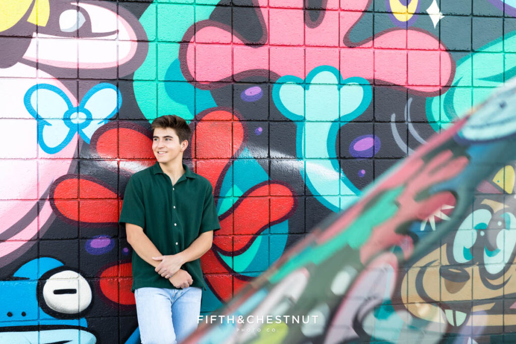 A senior wearing a casual green short sleeve button up and light washed jeans in front of a Hannah Eddy art installation in Midtown Reno for senior portraits by Reno Senior Photographer