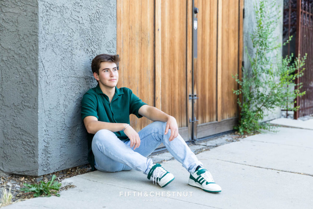 Casual and creative midtown senior photo of a senior boy wearing a green button up and light washed jeans in front of a stucco wall with a wooden door.