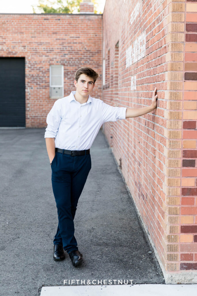 A senior wearing a white button up and navy slacks with black dress shoes poses for Creative Midtown Senior Portraits by Reno Senior Photographer against a brick building.