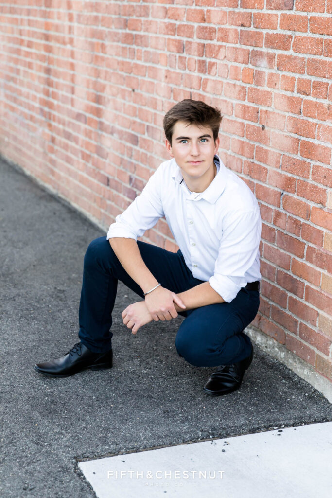 A senior wearing a white button up and navy slacks with black dress shoes poses for Creative Midtown Senior Portraits by Reno Senior Photographer against a brick building.