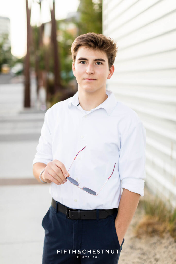 A senior wearing a white button up and navy slacks with black dress shoes poses for Creative Midtown Senior Portraits by Reno Senior Photographer against a metal wall while holding his aviator sunglasses and smiling.