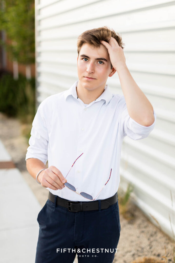 A senior wearing a white button up and navy slacks with black dress shoes poses for Creative Midtown Senior Portraits by Reno Senior Photographer against a metal wall while holding his aviator sunglasses.