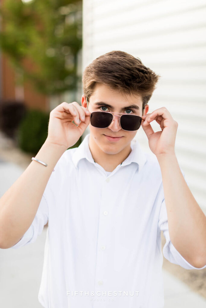 A senior wearing a white button up and navy slacks with black dress shoes poses for Creative Midtown Senior Portraits by Reno Senior Photographer against a metal wall while wearing his aviator sunglasses with his eyes in view.